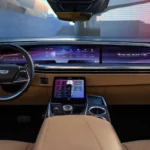 The driver's view of the 2026 Escalade IQL reveals the sweeping digital display with "ESCALADE IQL" branding and the elevated center console with climate and vehicle controls. The two-tone black and tan interior exemplifies Cadillac's premium craftsmanship.