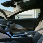 Interior view of the 2025 Lucid Air driver's cockpit featuring a two-spoke steering wheel with Lucid branding, floating curved digital display, wood trim accents, and light-colored upholstery in a minimalist luxury design.