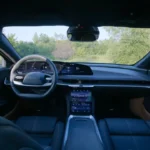 Interior view from the driver's perspective of the 2025 Lucid Air showing the dark blue interior with two-spoke steering wheel, curved digital display spanning the dashboard, center touchscreen, and silver trim accents with a view of trees through the windshield.