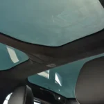 Interior view looking up at the 2025 Lucid Air's extensive glass roof panel with dark headliner material and support structure, showing a blue sky visible through the transparent roof from the interior of the vehicle.