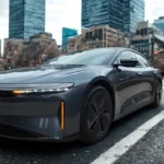 Side view of a gray 2025 Lucid Air electric sedan with distinctive aerodynamic styling and black wheels driving on a city street with modern buildings in the background during daytime.