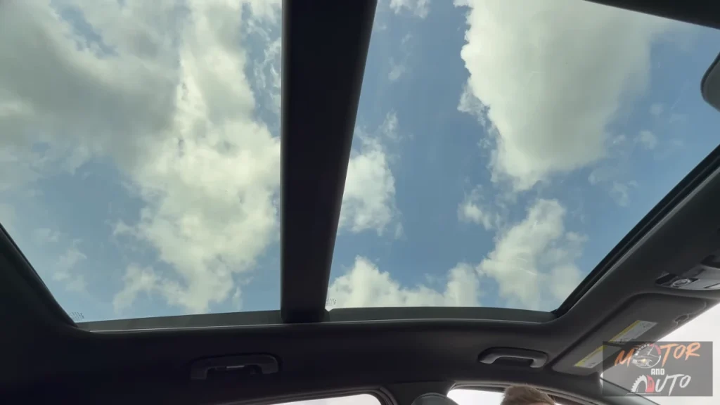 Interior view looking up through 2026 Kia Sportage panoramic sunroof showing blue sky with clouds and sunroof frame structure