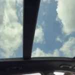 Interior view looking up through 2026 Kia Sportage panoramic sunroof showing blue sky with clouds and sunroof frame structure