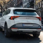 Rear three-quarter view of white 2026 Kia Sportage showing LED taillights, spoiler with hidden wiper, Sportage badging, and urban street environment