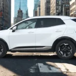 Side view of white 2026 Kia Sportage showing SUV proportions, black body cladding, alloy wheels, and urban street setting with city buildings