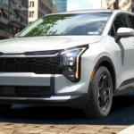 White 2026 Kia Sportage front three-quarter view with LED headlights illuminated showing amber DRL, modern front design, and urban street setting